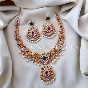 Neelkanthi Peacock Glory necklace set with twin peacock motifs, emerald-green and ruby stones, matte gold finish, and pearl drop detailing.