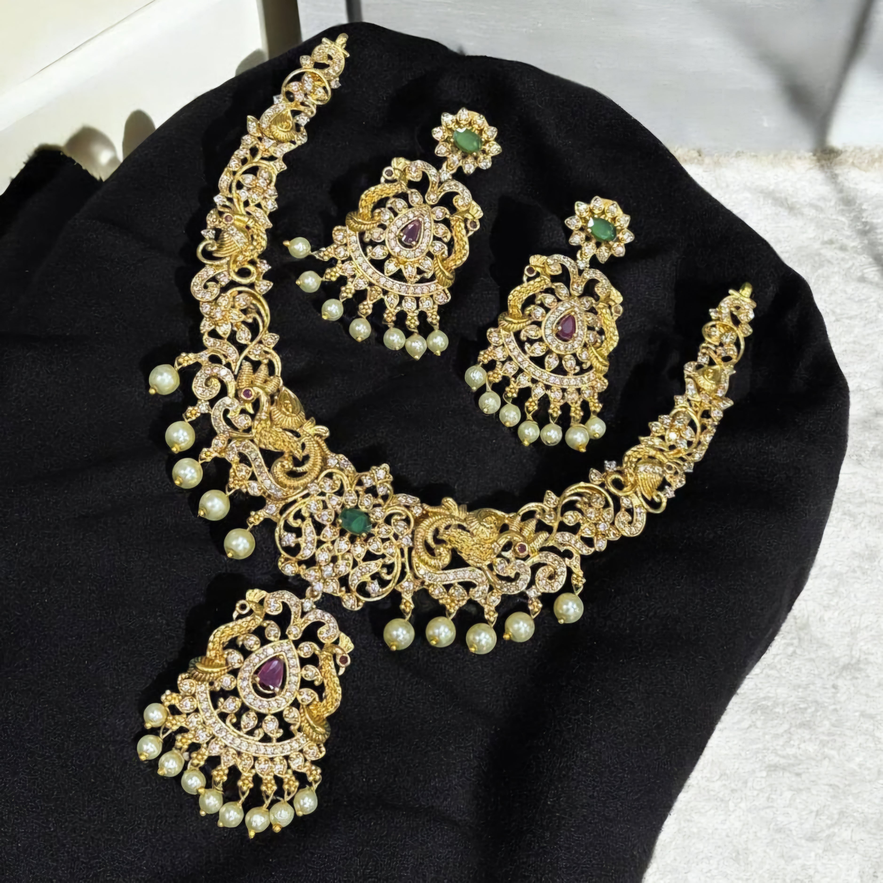 Neelkanthi Peacock Glory necklace set with twin peacock motifs, emerald-green and ruby stones, matte gold finish, and pearl drop detailing.