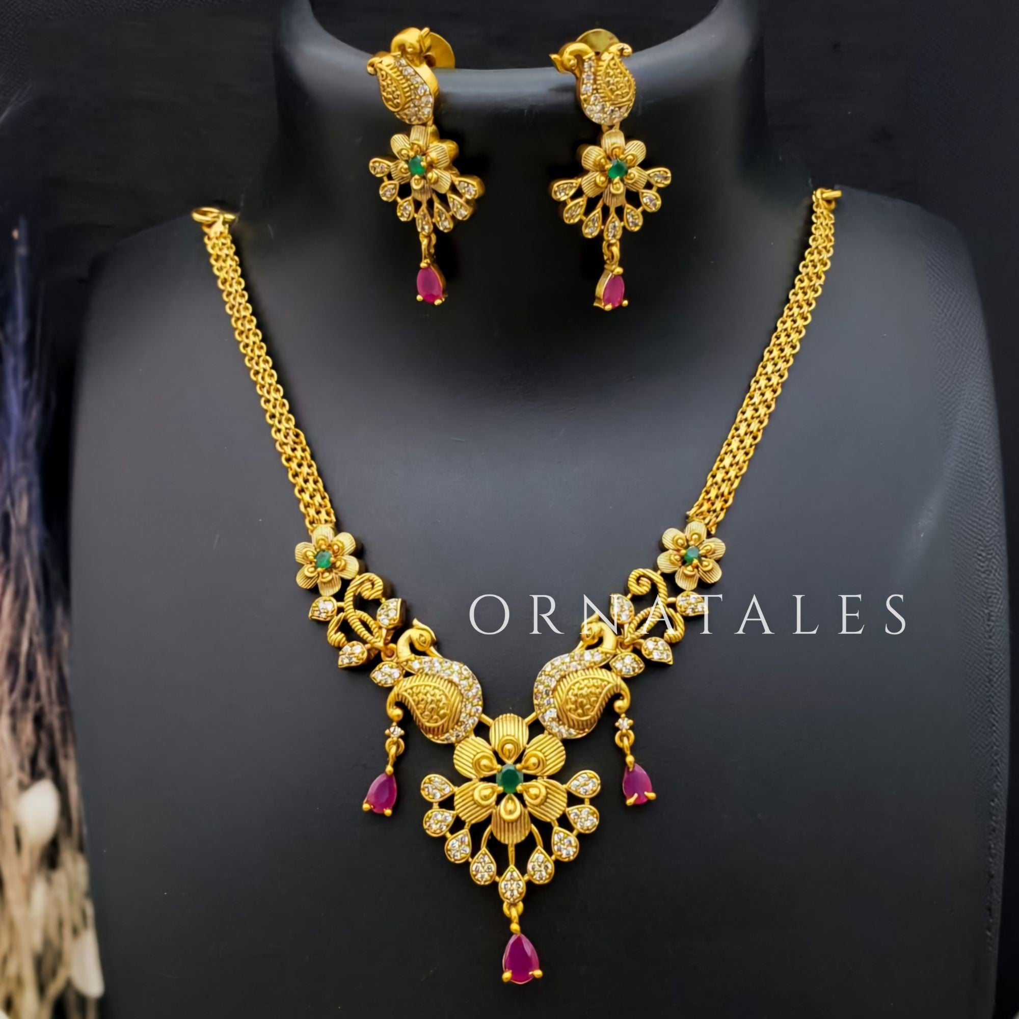 Temple-inspired peacock motif necklace with ruby and emerald stones, teardrop pendant, pearl drops, and antique gold finish.