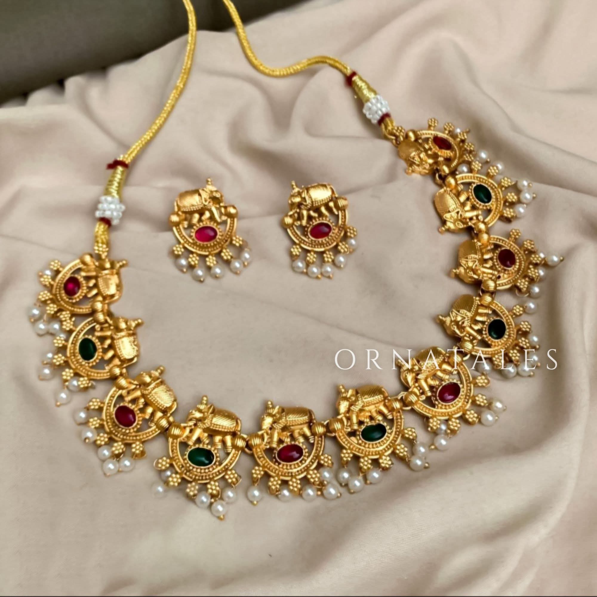 Matte gold South Indian choker with temple-style elephant (Hatimala) motifs, red and green stones, pearl fringe, and matching jhumka earrings.