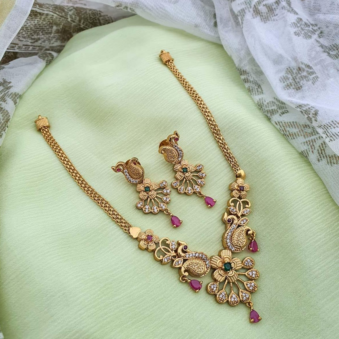 Temple-inspired peacock motif necklace with ruby and emerald stones, teardrop pendant, pearl drops, and antique gold finish.