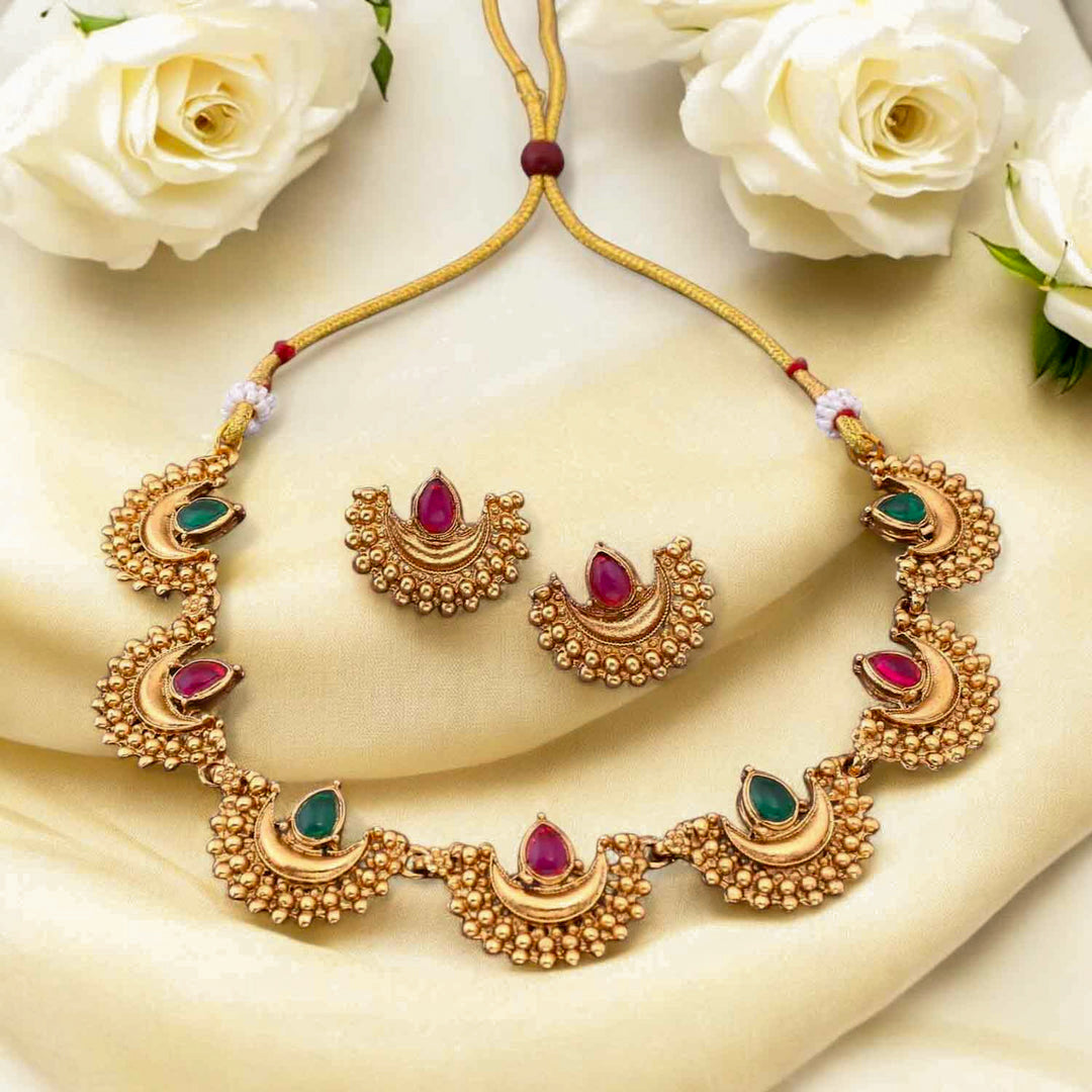South Indian choker necklace with crescent motifs, red and green teardrop stones, antique matte gold finish, and matching half-moon earrings.